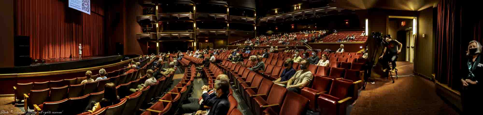 Spaced, assigned seating with masks and limited to 200. Total seating capacity is 1,341 divided into Orchestra (directly in front of camera), Tier (right at back), Balcony (above tier) and Box (directly above camera and in the same arrangement directly across). Emily Behrmann, director Midwest Trust Center (formerly Carlsen Center ) at JCCC is giving the welcoming speech and introduction to the show. One of the four camera positions is next to the doorway at right, paired with one on the other side, one in the pit moving left and right on dolly tracks and one at back of the house high near control booth. - Photo, Copyright 2021 Mike Strong all rights reserved